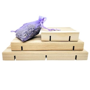 May include: A stack of three unfinished wooden soap dishes with a purple mesh bag filled with lavender buds. The soap dishes have a natural wood grain and a simple, rectangular design with cut-out slots.