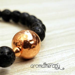 May include: A black lava rock bracelet with a hammered copper bead. The bracelet is on a light wood surface. "aromatherapy" is written in cursive on the surface.