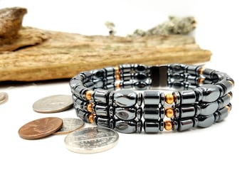 May include: A triple-strand bracelet made of black hematite beads with copper accents. The bracelet has a magnetic clasp.