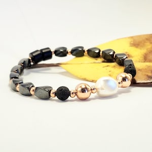 May include: A beaded bracelet featuring dark gray, rectangular hematite stones, interspersed with small copper-colored beads. The bracelet also includes black lava beads, a large white pearl, and copper-colored spherical beads. The bracelet is displayed on a white surface.