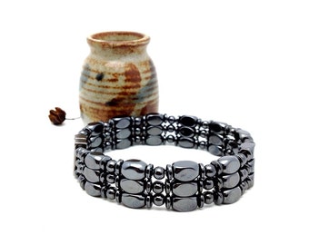 May include: A triple-strand bracelet composed of polished, dark gray, metallic-looking oval and spherical beads. The beads are arranged in three parallel rows, connected by a metallic clasp, creating a uniform, reflective, and smooth surface texture.