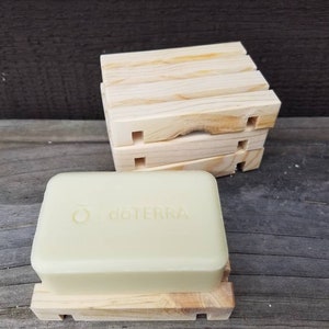 May include: A light-colored bar of soap with the "doTERRA" logo rests on a small, light-colored wooden soap dish. Several additional wooden soap dishes are stacked behind the soap. The soap and dishes are made of natural materials.