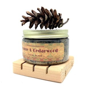 May include: A glass jar with a gold lid filled with a brown and white facial scrub. The jar has a label that reads "Orange & Cedarwood Facial Scrub 4 Ounces" and is sitting on a wooden soap dish. A pine cone is sitting on top of the jar.