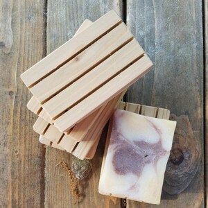 May include: A set of three wooden soap dishes with a natural finish. The dishes have grooves to help drain water and keep soap dry. A bar of handmade soap with swirls of white, brown, and pink is sitting on one of the dishes.