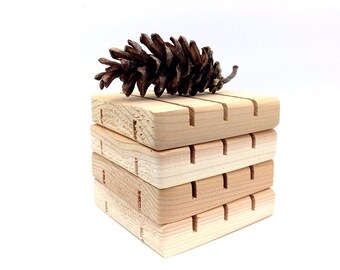 May include: A stack of four wooden soap dishes with grooves to help soap drain. The dishes are made of natural wood and have a light brown color. A pine cone sits on top of the stack.