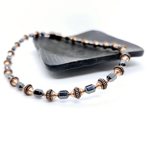 May include: A beaded bracelet featuring alternating dark gray and copper-colored beads. The bracelet is made of small, geometric-shaped dark gray beads and copper-colored, cylindrical beads. The bracelet is displayed on a dark gray, rectangular surface against a white background.