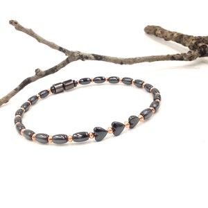 May include: A black beaded bracelet with copper-colored accents and heart-shaped beads. The bracelet is strung on a flexible cord and has a magnetic clasp. The bracelet is displayed on a white surface with a twig in the background.