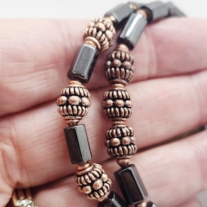 May include: A beaded bracelet featuring alternating dark gray cylindrical beads and round, textured copper-toned metal spacer beads. The bracelet is constructed with a repeating pattern of one cylindrical bead followed by one round metal bead.