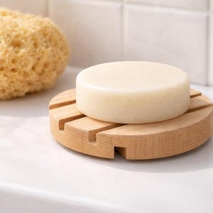 May include: A round, light-colored soap bar rests on a wooden soap dish with a slotted design. A natural sea sponge is in the background. The soap dish is made of light-colored wood and is on a white surface.