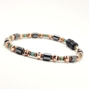May include: A beaded bracelet featuring a combination of dark gray, rose gold, and turquoise beads. The bracelet has a magnetic clasp and is displayed against a white background. The beads are arranged in a repeating pattern.