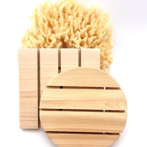May include: A natural sea sponge with a light yellow color rests above a wooden soap dish and a round wooden soap rest. The soap dish is rectangular with vertical slots, and the soap rest is circular with horizontal slots. The items are made of light-colored wood.