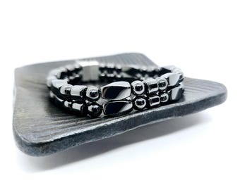 May include: A black magnetic bracelet with two rows of beads. The bracelet is made of round and oval-shaped beads.