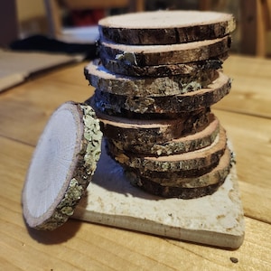 May include: A stack of rustic wooden coasters made from tree slices. The coasters have a natural bark edge and a light-colored wood center. One coaster is separate from the stack, resting on a small, square, off-white tile. The coasters are on a wooden surface.