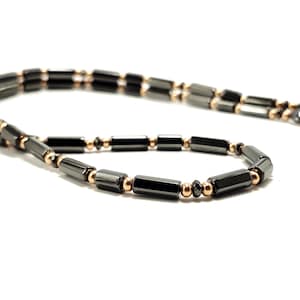 May include: A black and gold-toned necklace with a magnetic clasp. The necklace features rectangular black beads interspersed with small, round gold-colored beads. The jewelry is displayed against a white background.