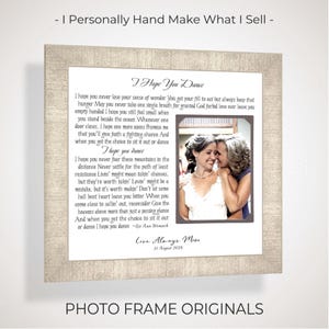 May include: A square framed print featuring the lyrics to I Hope You Dance by Lee Ann Womack, a photograph of two people embracing, and the text Love Always, Mom 31 August 2026. The frame has a light beige textured finish and a white mat border.