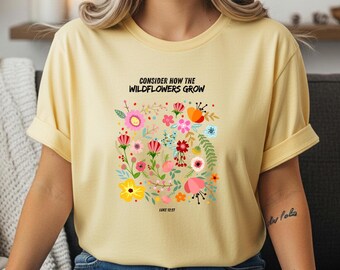 Puede incluir: Una camiseta de manga corta de color amarillo claro con un gráfico colorido de varias flores silvestres y el texto negro "CONSIDER HOW THE WILDFLOWERS GROW" sobre las flores y "LUKE 12:27" debajo del diseño floral en la parte delantera.