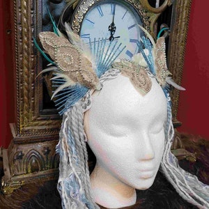 May include: A decorative headpiece with a white base, adorned with beaded embellishments, feathers, and blue accents. The headpiece is displayed on a mannequin head with long, gray and blue braided extensions. The background includes a vintage clock.