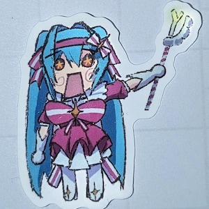 Può includere: Un adesivo con un personaggio di cartoni animati in stile anime con capelli blu, un abito rosa e bianco e un bastone. Il personaggio ha grandi occhi arancioni e la bocca aperta, che esprime sorpresa. L'adesivo ha un bordo bianco.