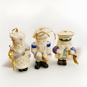 May include: Three ceramic Christmas ornaments in the shape of figures. The ornaments are white with gold accents, and each has a different design. One holds a staff, and the others have hats with the years 2001, 2003, and 2006.