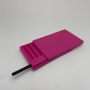 Peut inclure: Une boîte rectangulaire rose vif avec un couvercle coulissant et trois compartiments encastrés. Une tige cylindrique noire dépasse du côté. La boîte est faite d'un matériau solide et lisse.