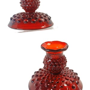 May include: Two red glass candle holders with a hobnail design. The candle holders have a round base and a flared top. The glass is a deep red color and has a glossy finish. The candle holders are on a white background.