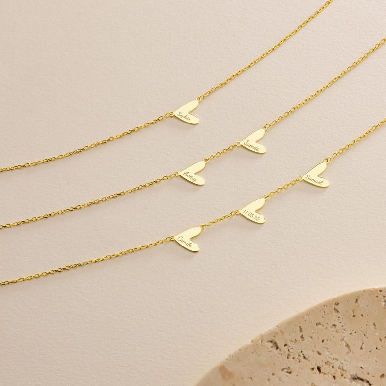 May include: Gold-colored necklaces with heart-shaped pendants. Each heart pendant has a name or date engraved on it. The necklaces are displayed on a light-colored surface.