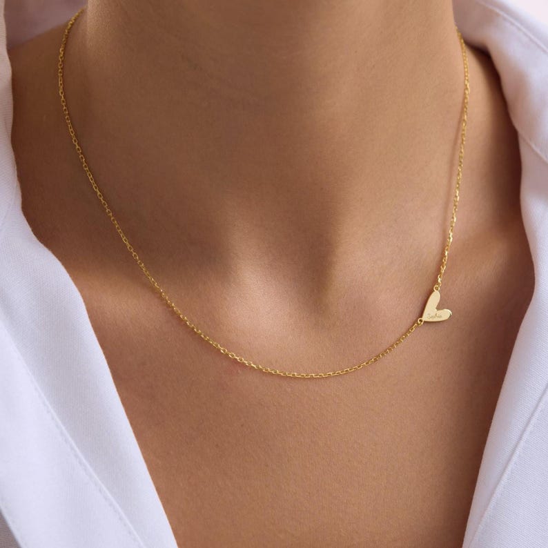 May include: A delicate gold necklace featuring a small heart pendant. The heart is positioned to the side of the chain. The necklace is worn against a white shirt.