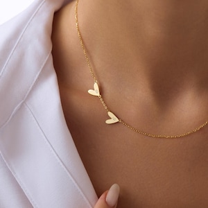 May include: A delicate gold necklace featuring two heart pendants. Each heart has a single initial engraved on it. The necklace is worn against a white shirt.
