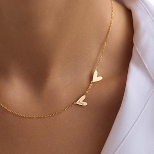 May include: A gold-colored necklace featuring three heart-shaped pendants. One pendant is inscribed with the name "Avery". The necklace is worn against a white shirt.