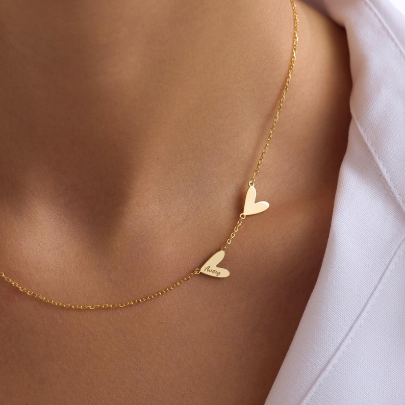 May include: A gold-colored necklace featuring three heart-shaped pendants. One pendant is inscribed with the name "Avery". The necklace is worn against a white shirt.