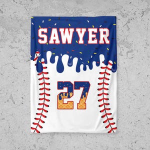 May include: A white baseball-themed blanket with a blue top section featuring the name "SAWYER" in white block letters. The number "27" is in the center, with a waffle-like texture. Red stitching lines the sides.