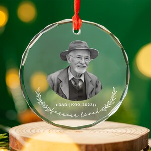Può includere: Un ornamento natalizio in vetro trasparente e sfaccettato con la foto di un uomo anziano con cappello e occhiali. L'ornamento presenta la scritta "+ DAD (1939-2022) + forever loved" e un nastro rosso per appenderlo.