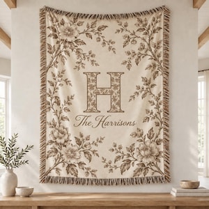 May include: A cream-coloured woven throw blanket featuring a large brown letter H monogram and the text The Harrisons. The blanket is decorated with brown floral vine patterns along the edges and has a fringed border on the top and bottom.