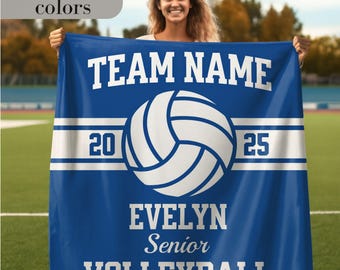 May include: A blue volleyball blanket with white text and a volleyball graphic. The blanket features the text "TEAM NAME", "20", "25", "EVELYN", "Senior", and "VOLLEYBALL". The blanket is held by a person in a field.