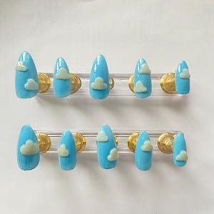 May include: Ten artificial nails, each painted a bright sky blue. Each nail features a small, light green cloud design. The nails are arranged in two rows of five, mounted on clear plastic holders with gold-coloured accents.