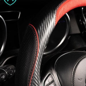 May include: Close-up of a black and red steering wheel cover with a carbon fiber texture. The cover has red stitching and a perforated grip area. Text on the image reads "ANTI-SLIP GRIP & ERGONOMIC COMFORT ZONE" and "Textured Design Ensures Confident Grip."
