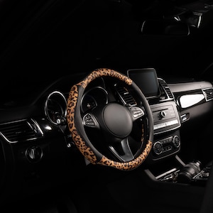 May include: A steering wheel cover featuring a brown and black leopard print pattern, installed on a black car steering wheel. The cover has a textured surface and is positioned within the interior of a vehicle with a dark dashboard and console.
