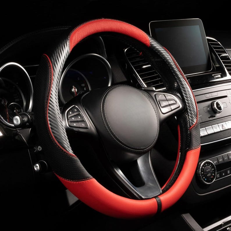 May include: A black and red steering wheel cover. The cover has a carbon fiber texture on the sides and a smooth red finish on the top and bottom. The steering wheel is in a car interior.