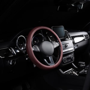 May include: A dark maroon steering wheel cover with a textured, perforated pattern installed on a black vehicle steering wheel. The cover features a smooth outer rim and a grip-enhancing design, contrasting against the dark interior dashboard.