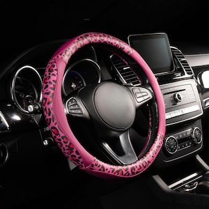 Pode incluir: Uma capa de volante rosa com estampa de leopardo preto instalada em um carro. A capa é rosa vibrante com um padrão de estampa de leopardo preto. O interior do carro é preto, com uma tela digital e um painel de controle.