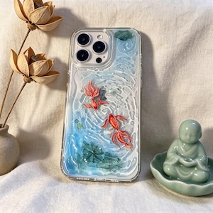 Könnte beinhalten: Transparente Handyhülle mit einem Design von zwei roten Goldfischen, die in blauem Wasser mit Seerosen schwimmen. Die Hülle hat einen transparenten Rand und ein glänzendes Finish. Das Design ist künstlerisch und detailliert.