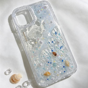Può includere: Custodia per telefono trasparente con un design liquido e glitterato, con conchiglie, perle e piccoli accenti blu e gialli. La custodia ha un bordo trasparente con un bordo perlato.