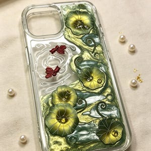 May include: Transparent phone case with a decorative design featuring green and gold lily pads, red dragonflies, and a pearlescent background. The case has a clear border and is embellished with small pearls.