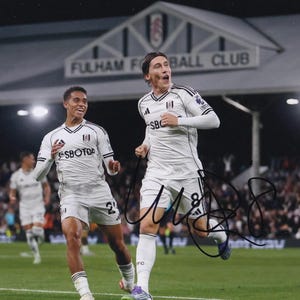 Op de afbeelding: Twee voetballers in witte en zwarte tenues vieren op een groen veld. Het bord van Fulham Football Club is zichtbaar op de achtergrond. De spelers dragen witte shorts en shirts met het Fulham FC-logo.