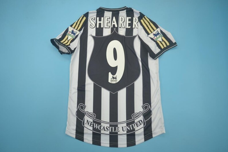 May include: A black and white striped Newcastle United soccer jersey featuring the name SHEARER and the number 9 on the back. The shirt includes a Premier League patch on each sleeve and a banner graphic at the bottom with the text NEWCASTLE UNITED.