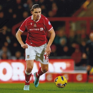 May include: A soccer player in a red jersey with white shorts and red socks, dribbling a yellow and blue soccer ball on a green field. The jersey has the word "UNITED" on it. The background shows a crowd of people.