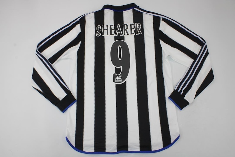 May include: A long-sleeve soccer jersey featuring vertical black and white stripes. The back displays the name SHEARER in white letters above a large black number 9. A small Premier League lion emblem is printed below the number 9.