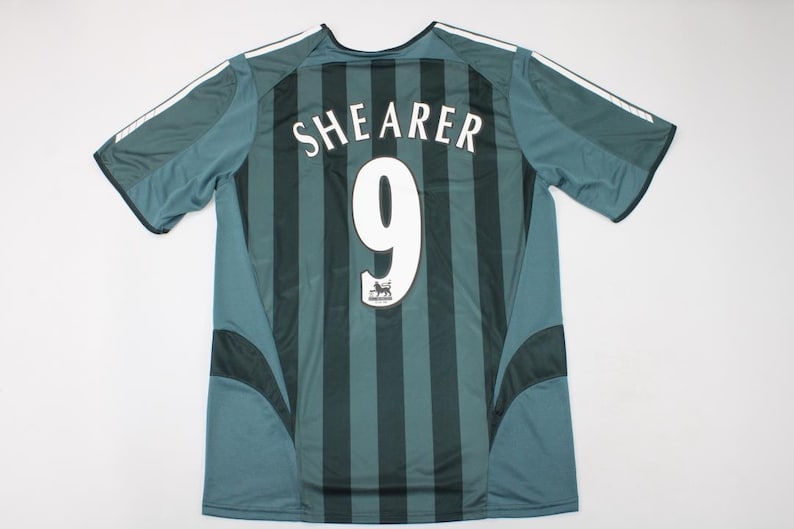 Puede incluir: Una camiseta deportiva de manga corta con rayas verticales de color verde oscuro y verde azulado. La parte posterior muestra el nombre SHEARER en letras blancas sobre el n&uacute;mero 9 en blanco, con un peque&ntilde;o emblema de le&oacute;n debajo del n&uacute;mero.