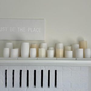 以下が含まれることがあります： 白とクリーム色の様々な高さと幅の円柱キャンドルのコレクションが白い棚に配置されています。棚の上には「JUST BE THE PLACE」と書かれた白いサインがあります。背景は白い壁と白いレンガの暖炉です。