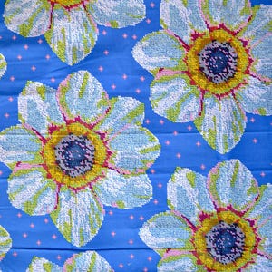 May include: A blue fabric with a repeating floral pattern. The flowers have light blue petals, yellow centers, and pink and green accents. Small pink dots are scattered across the blue background.